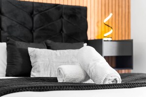 A neatly made bed with a black headboard, white pillows, and a rolled white towel on a dark, textured bedspread. - Luxury Two Bedroom Apartment - Jewellery Quarter (Birmingham)