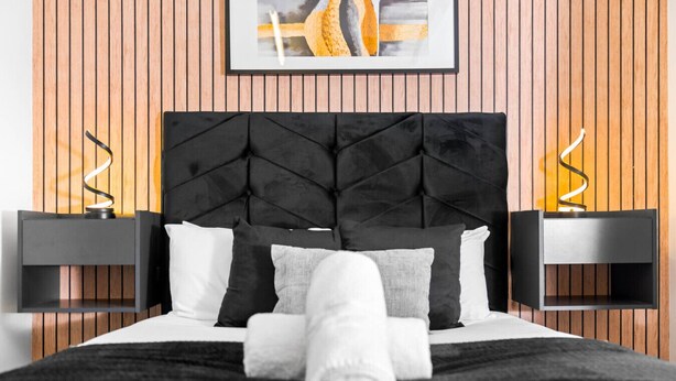 Ein modernes Schlafzimmer mit einer gepolsterten Kopfteil, einem ordentlich bezogenen Bett mit einem zusammengerollten Handtuch und zwei Nachttischen mit Wandlampen.