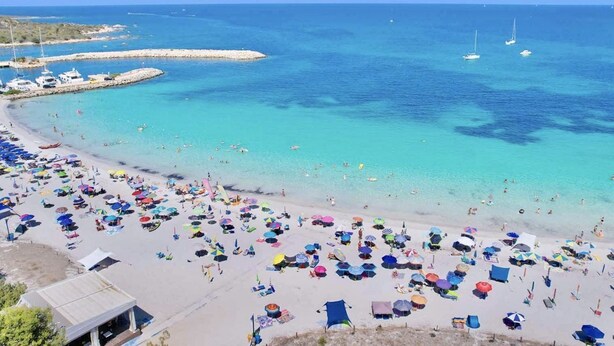 A beach with numerous colorful umbrellas and sun loungers, a clear blue sea, and boats in the distance.