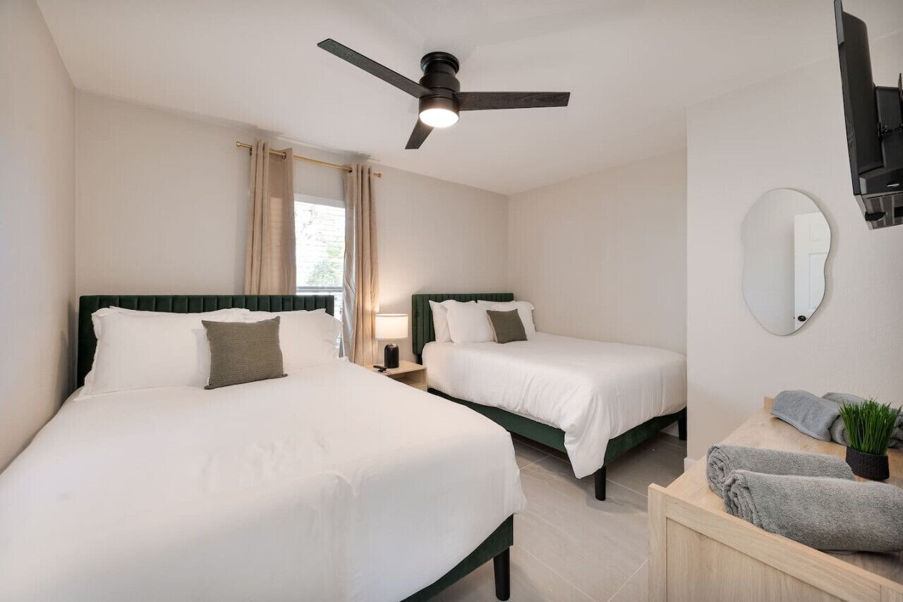 Quarto de hotel com duas camas, ventoinha no teto, televisão, espelho e uma mesinha com toalhas e uma planta.
