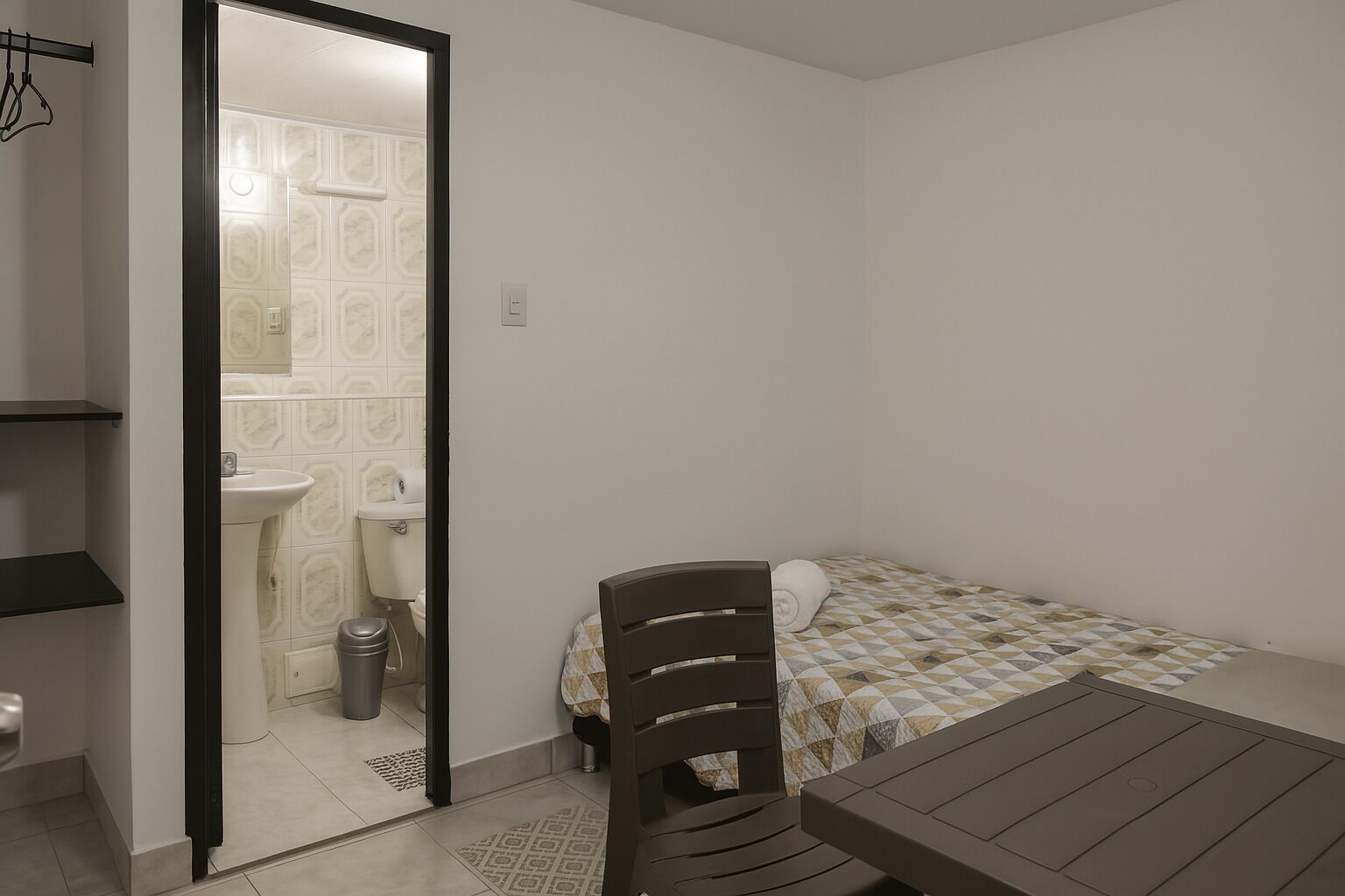 Une petite pièce, de taille modeste, comprenant un lit, une table et une chaise. On peut apercevoir une salle de bain à travers une porte ouverte, qui contient des toilettes, un lavabo et une poubelle.