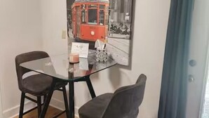 A dining area with a glass table and two chairs, a framed black and white photo of a tram on the wall, and a window with a curtain. - Charming studio in delightful Spring Branch for a quick, tranquil stay.   (Spring Branch)