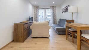 Ein gemütlicher Wohnraum mit einer Couch, einem Holztisch, einem Fernseher auf einer Kommode und Blick auf Gebäude durch ein Fenster mit floralem Vorhang.