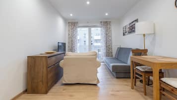 Un espacio habitable pequeño con un sofá, una mesa de madera, un televisor en un mueble, y vistas de edificios a través de una ventana con cortinas florales.