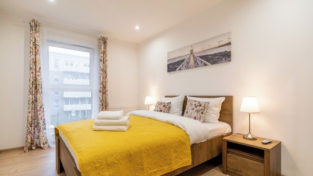 A hotel room with a bed, a nightstand, a lamp, a painting, and a window with curtains. - Park Śląski Homely Apartment #16 (Katowice)