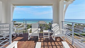 Une terrasse en bois avec des chaises blanches et une table, donnant sur une plage et l’océan.