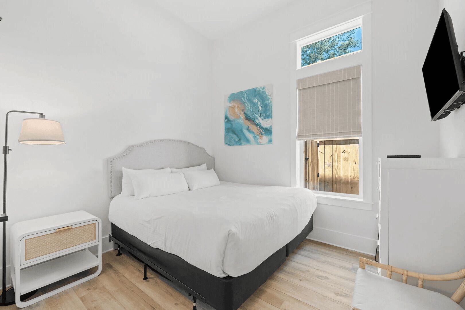 A bedroom with a bed, a bedside table, a lamp, a window, and a painting.