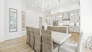 Uma ampla área de jantar com uma mesa comprida e cadeiras de vime, um lustre e uma cozinha moderna ao fundo.
