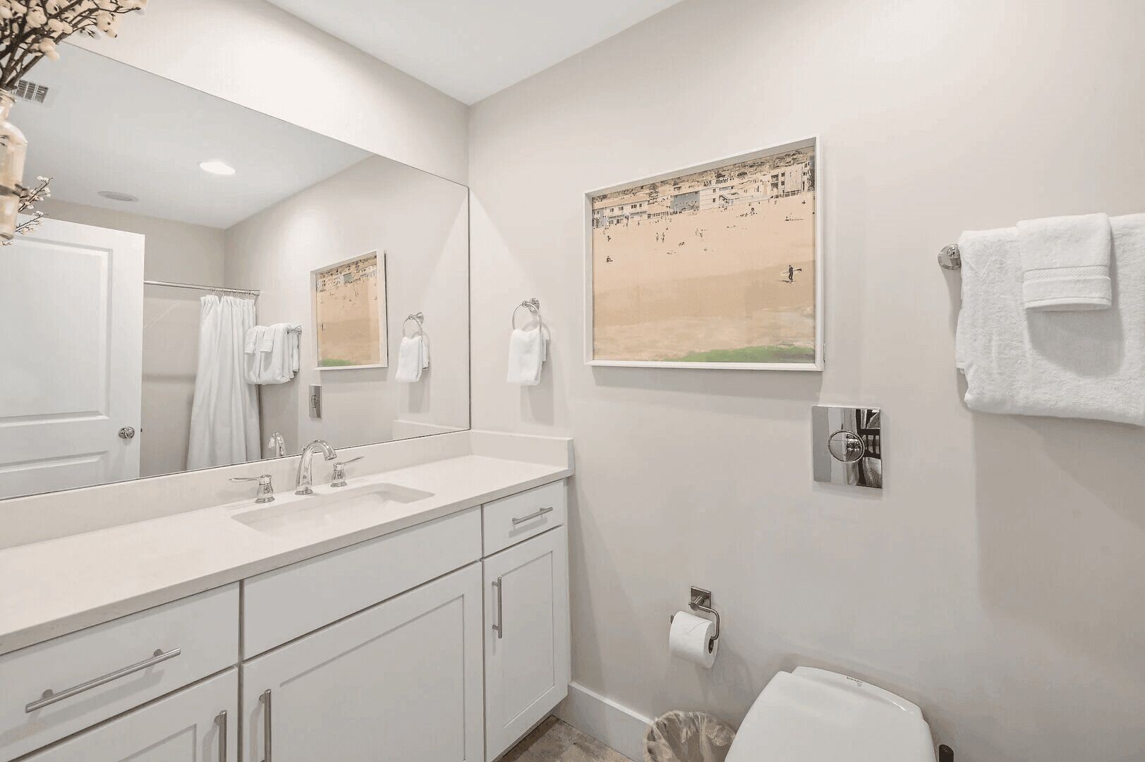 A modern bathroom with a large mirror, white fixtures, a sink, and a toilet. There is a framed picture on the wall and a towel rack with towels.