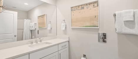 Een moderne badkamer met een grote spiegel, witte sanitair, een wastafel en een toilet. Er hangt een lijst met een foto aan de muur en een handdoekrek met handdoeken.