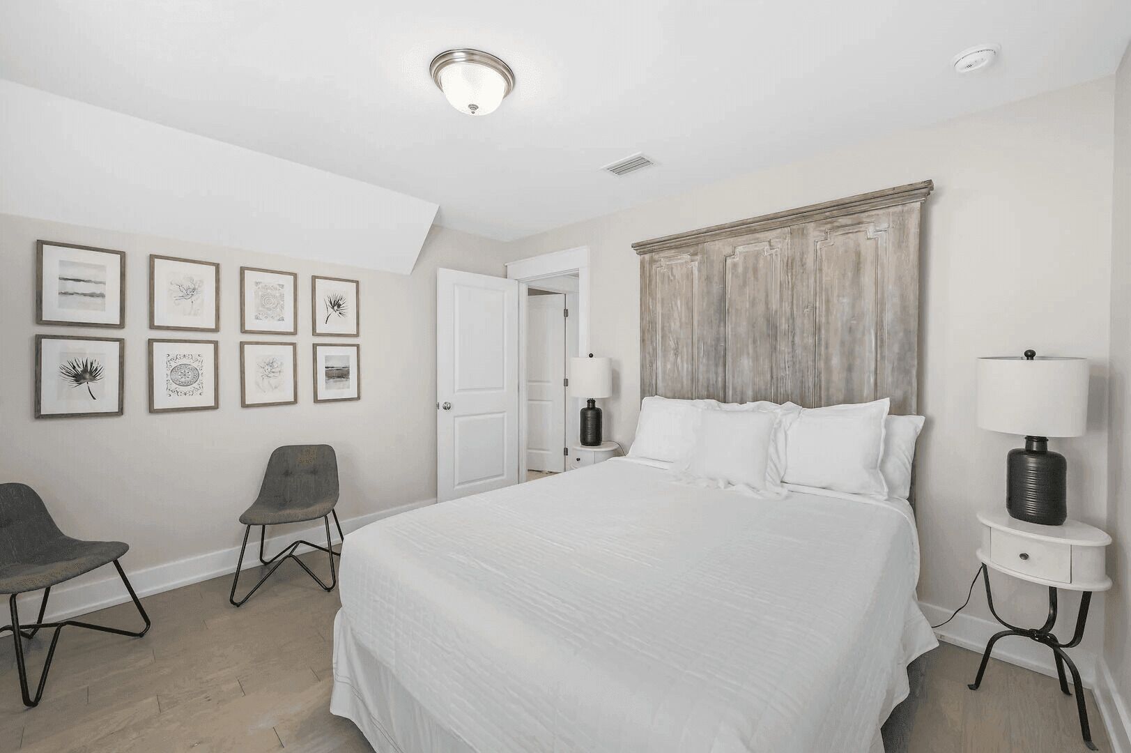 Un dormitorio con una cama grande, una silla, una mesita de noche con lámpara y cuadros en la pared.