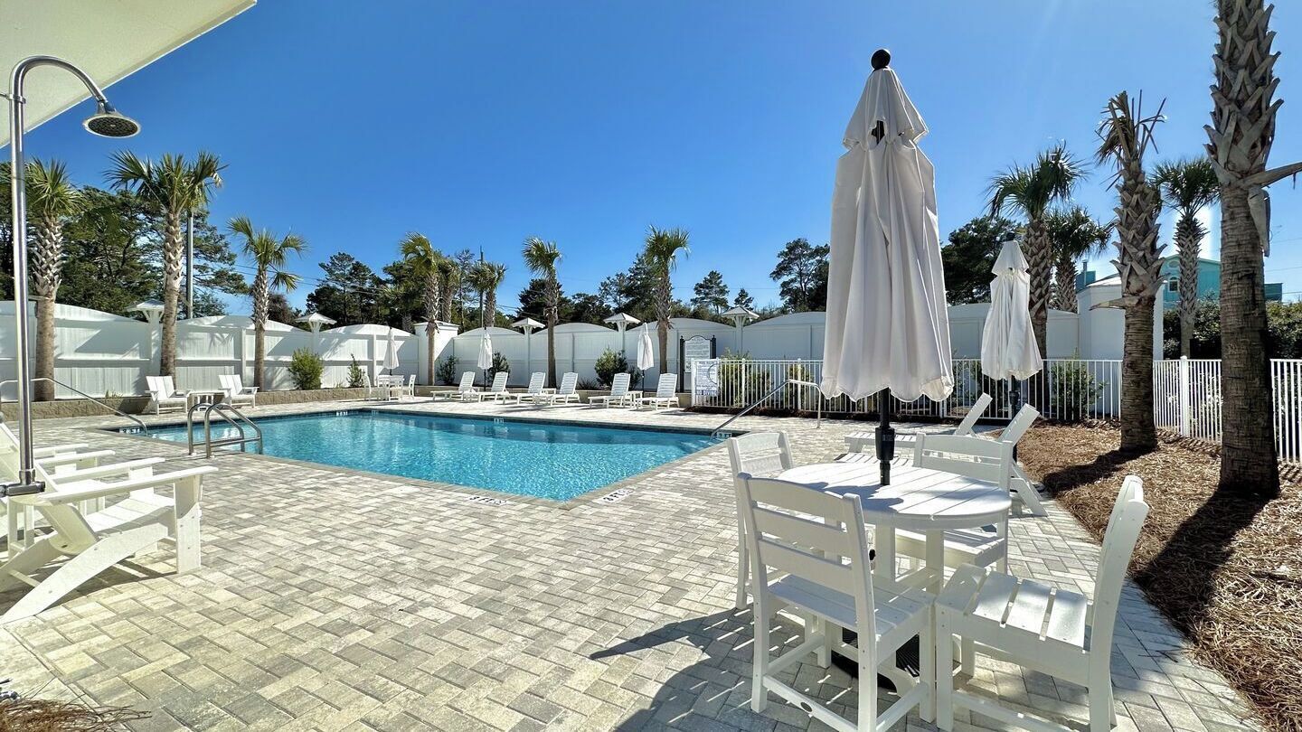Une piscine entourée de chaises longues et de parasols, bordée de palmiers et surplombée d’un ciel dégagé.