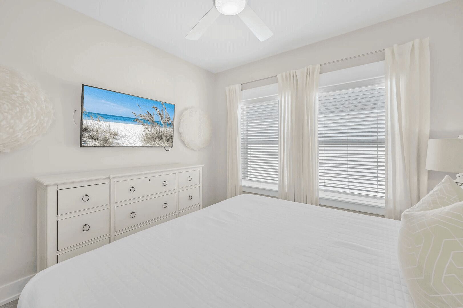 A bedroom with a large bed, a dresser with drawers, a ceiling fan, and a window with blinds.