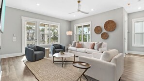 A spacious living room with a white sofa, two armchairs, a coffee table, and a ceiling fan. Large windows provide natural light and views of the outside. - Sea Dweller by Avantstay Rooftop Spa + Gulf View Golf Cart, Pool (Panama City Beach)