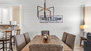 A dining area with a wooden table, four upholstered chairs, a chandelier, and a 'BEACH' sign on the wall.