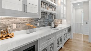 A modern kitchen with white cabinetry, a marble countertop, and a stone backsplash. There are glass jars with wooden lids on the counter, and a coffee machine.