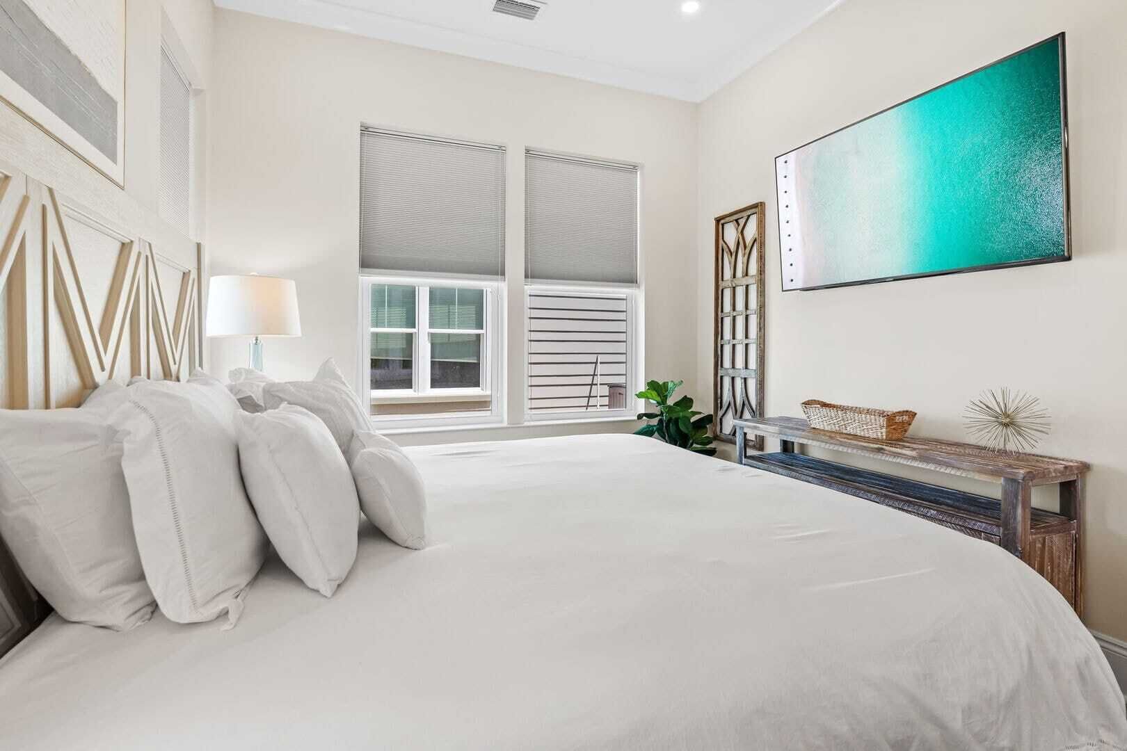 A bedroom with a large bed, a window with blinds, a painting, and a small table with a basket.