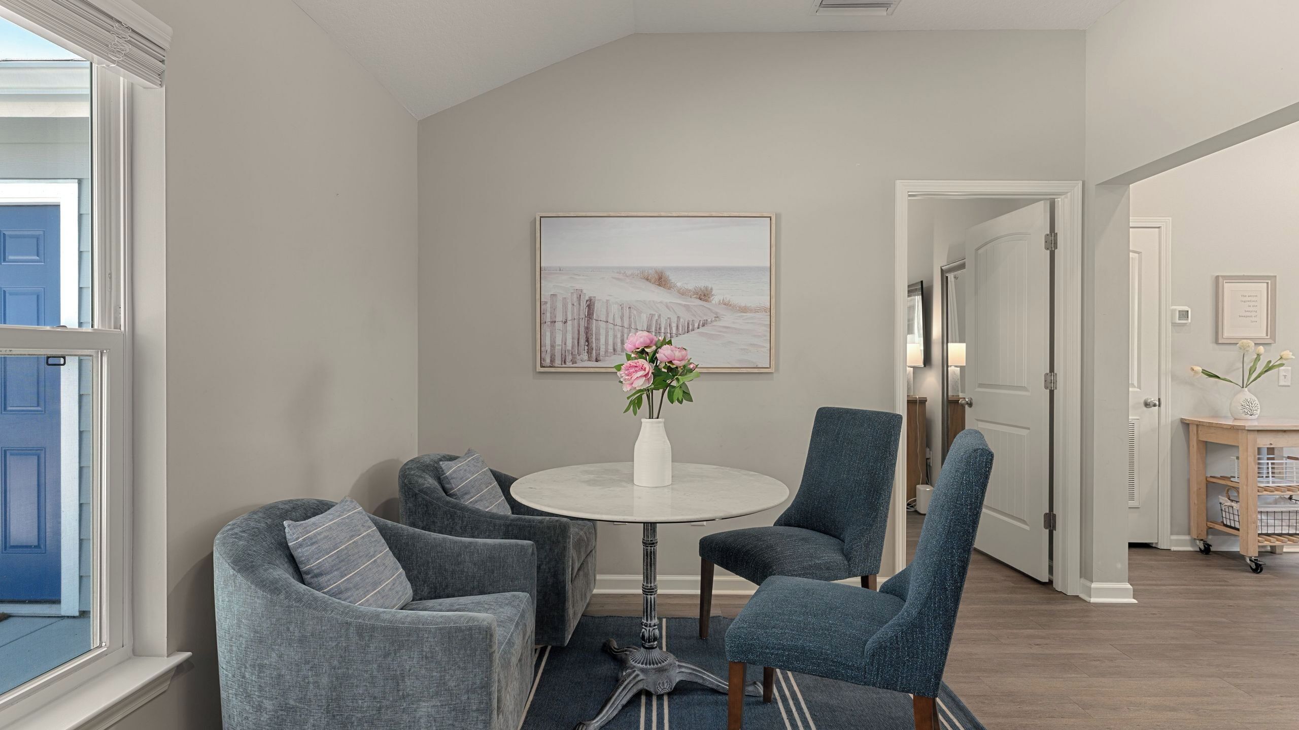 A room with a round table, two armchairs, a vase with flowers, a painting on the wall, and a door leading to another room.