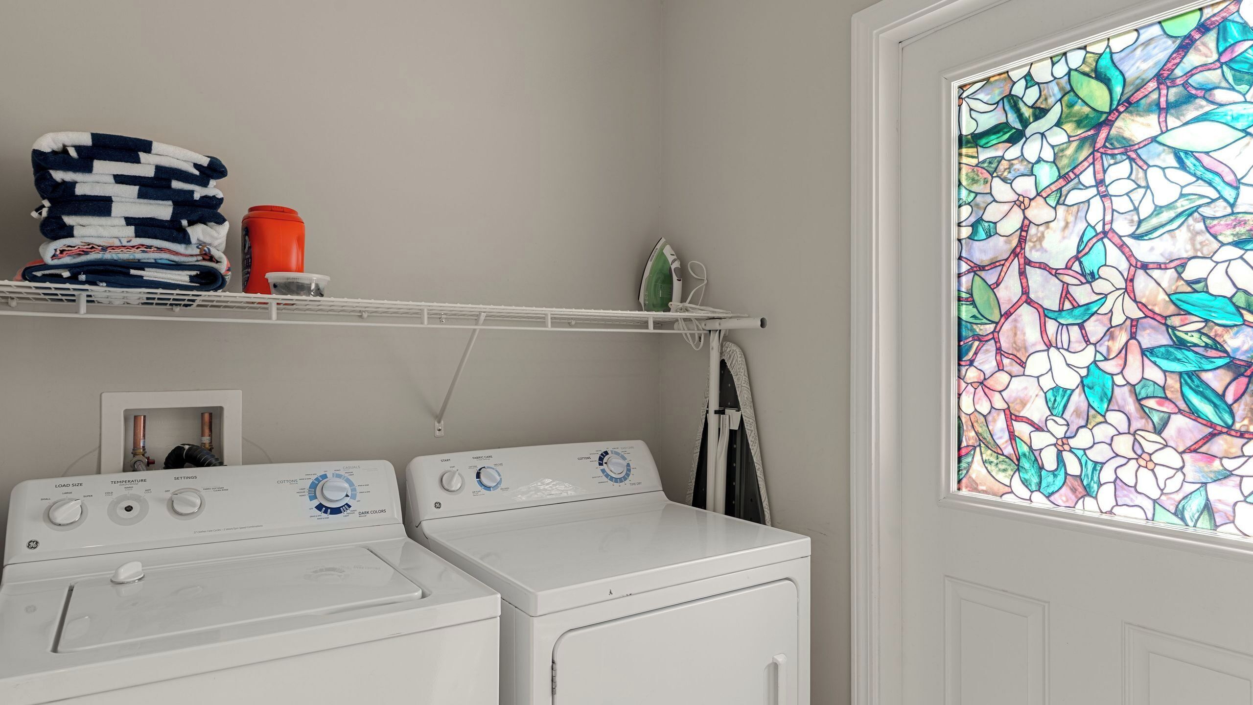 A laundry room with two washers and dryers, a shelf with folded towels, a red container, and a stained glass window.
