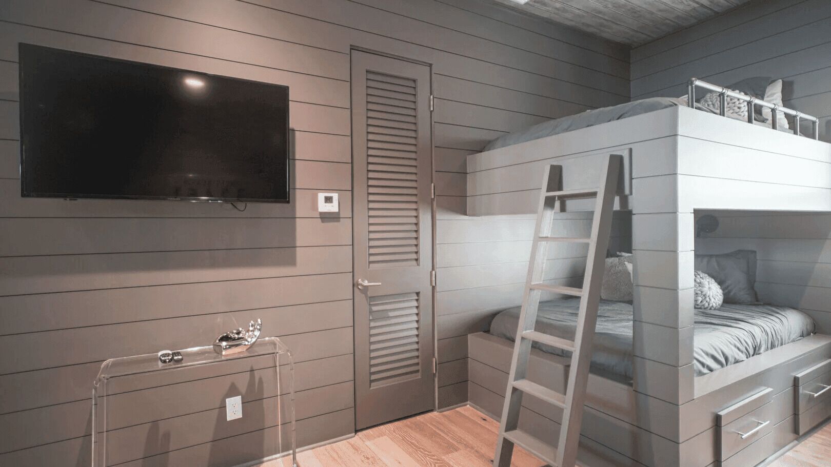 Un dormitorio con literas, un televisor montado en la pared, una puerta con ventana y una mesita con lámpara.
