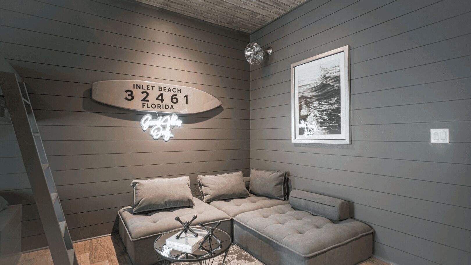 A room with a sectional sofa, a small round table, a neon sign, and a framed picture on the wall.