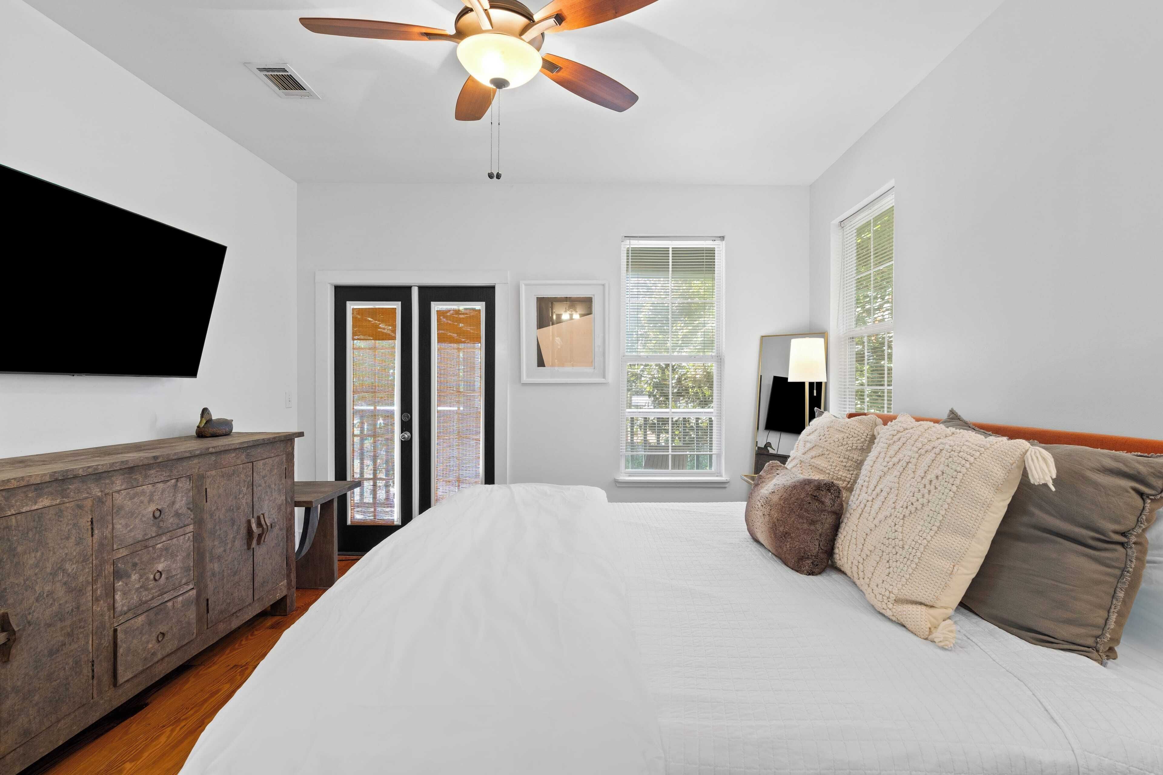 A bedroom with a bed, wooden dresser, ceiling fan, and large windows.
