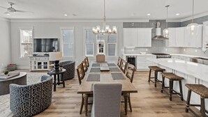 A modern kitchen with white cabinets, a central island, and a dining area with a wooden table and chairs. - 30A Salt Life by Avantstay Golf Cart + Pool Blue Mountain (Santa Rosa Beach)