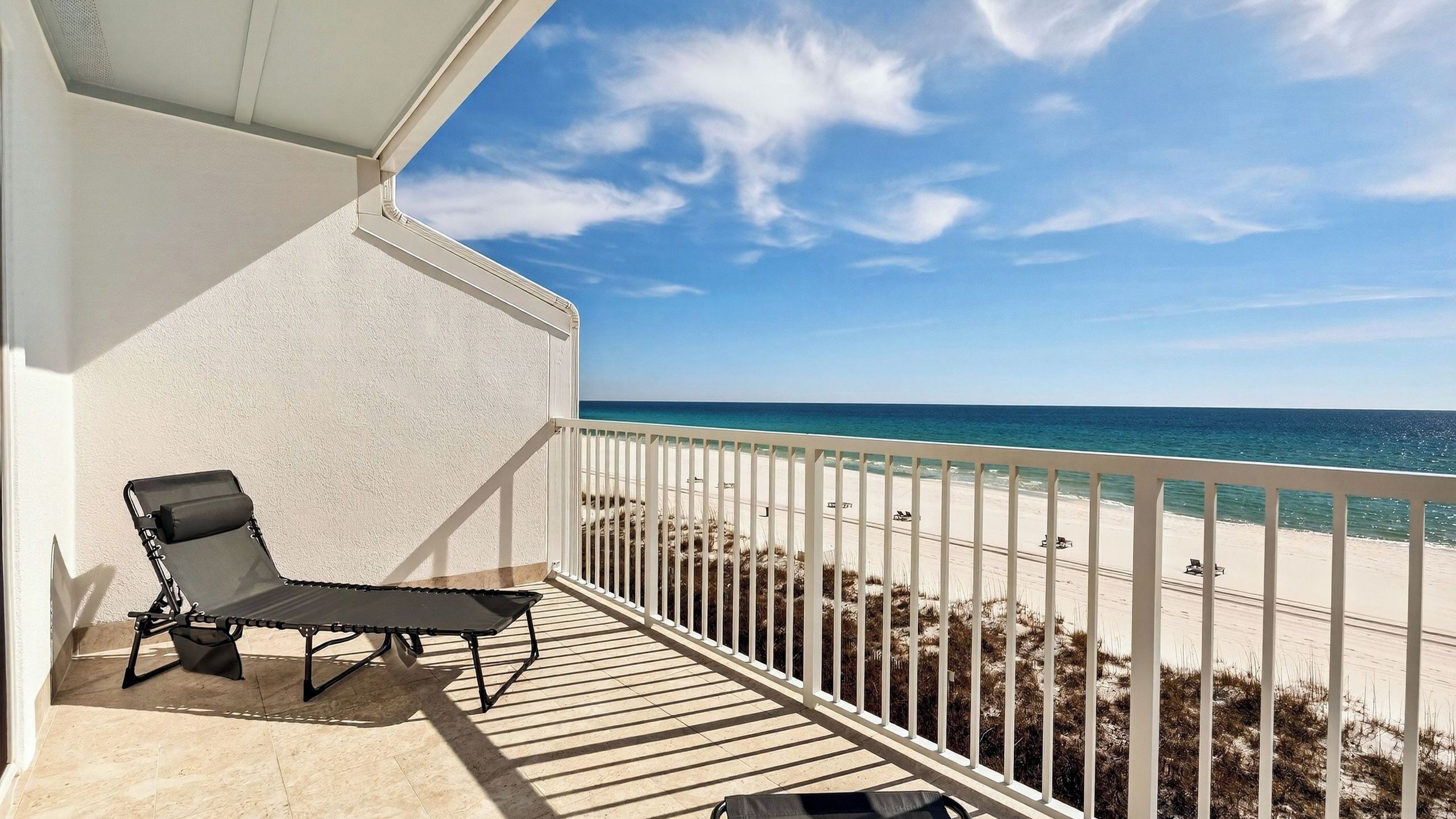 A balcony with a black lounge chair, a white railing, and a view of the beach and ocean.