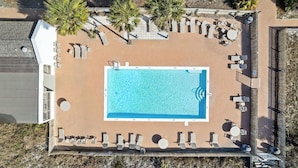 En vy från ovan av en rektangulär swimmingpool omgiven av solstolar och vilfåtöljer, med en stenlagd terrass och en byggnad på ena sidan.