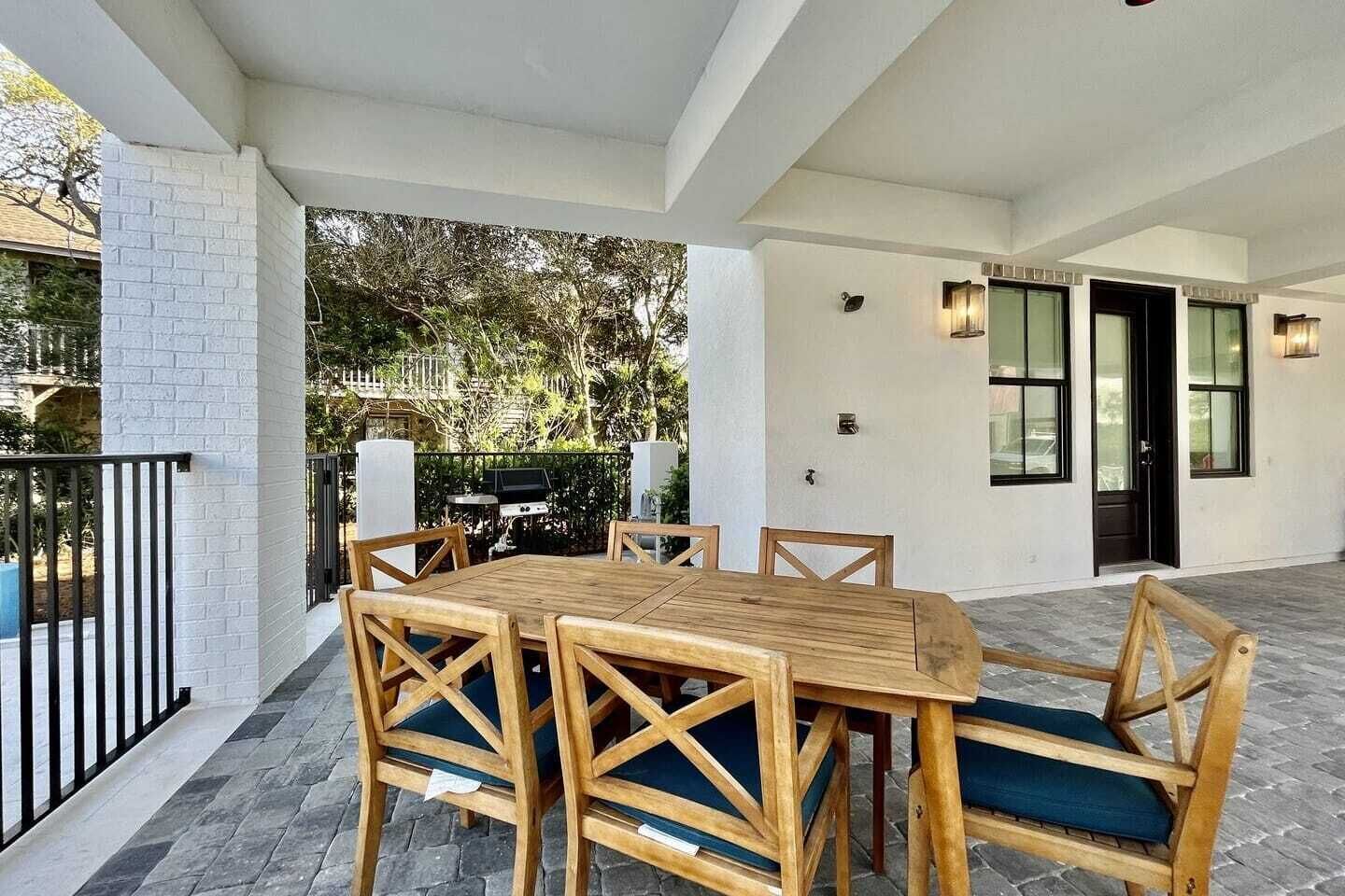 Une terrasse extérieure avec une table et des chaises en bois, un barbecue et un mur blanc agrémenté de fenêtres.