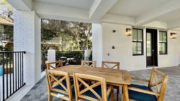Une terrasse extérieure avec une table et des chaises en bois, un barbecue et un mur blanc agrémenté de fenêtres.
