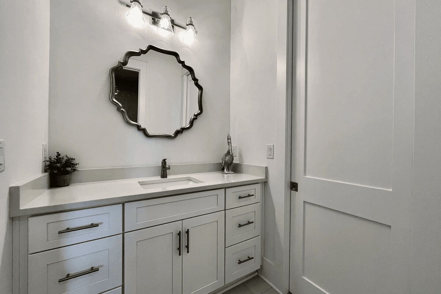 Une salle de bain avec un meuble-lavabo blanc, un miroir doté d’un cadre original et un luminaire placé au-dessus du miroir.