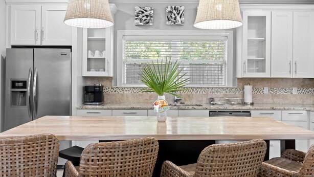 A modern kitchen with a large island, wicker chairs, stainless steel appliances, and pendant lighting.