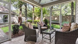 A covered patio with wicker furniture and a glass table, surrounded by a brick floor and large windows.