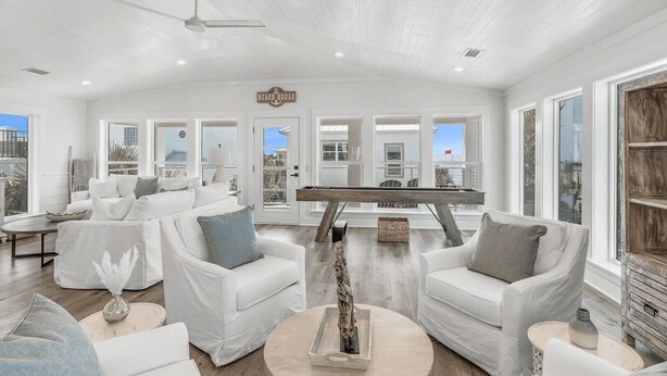 A spacious living room with a pool table, white sofas, and large windows offering a view of the beach.