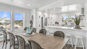 A modern kitchen with white cabinetry, a wooden dining table, wicker chairs, and a view of the outside.