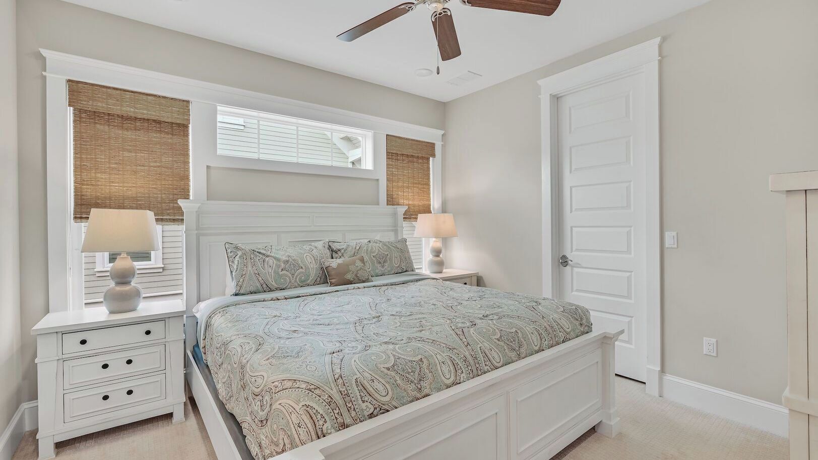 A bedroom with a bed, nightstands, lamps, a ceiling fan, and a door.