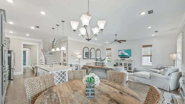 Amplia sala de estar con una mesa redonda de madera para comer, una cocina con armarios blancos y una escalera que conduce a un nivel superior.