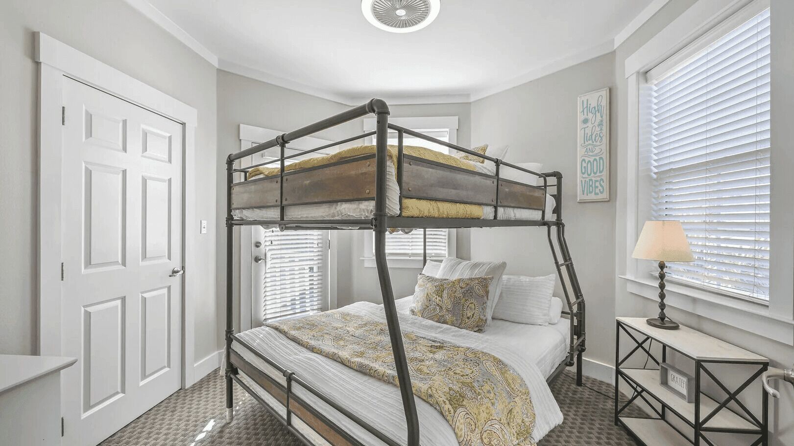 Una cama litera con cabecera, una mesita de noche con lámpara y una ventana con persianas.