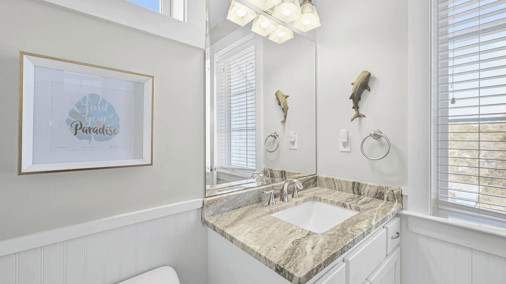 Une salle de bain avec un lavabo en marbre, une citation inspirante encadrée, un grand miroir et deux porte-serviettes fixés au mur.