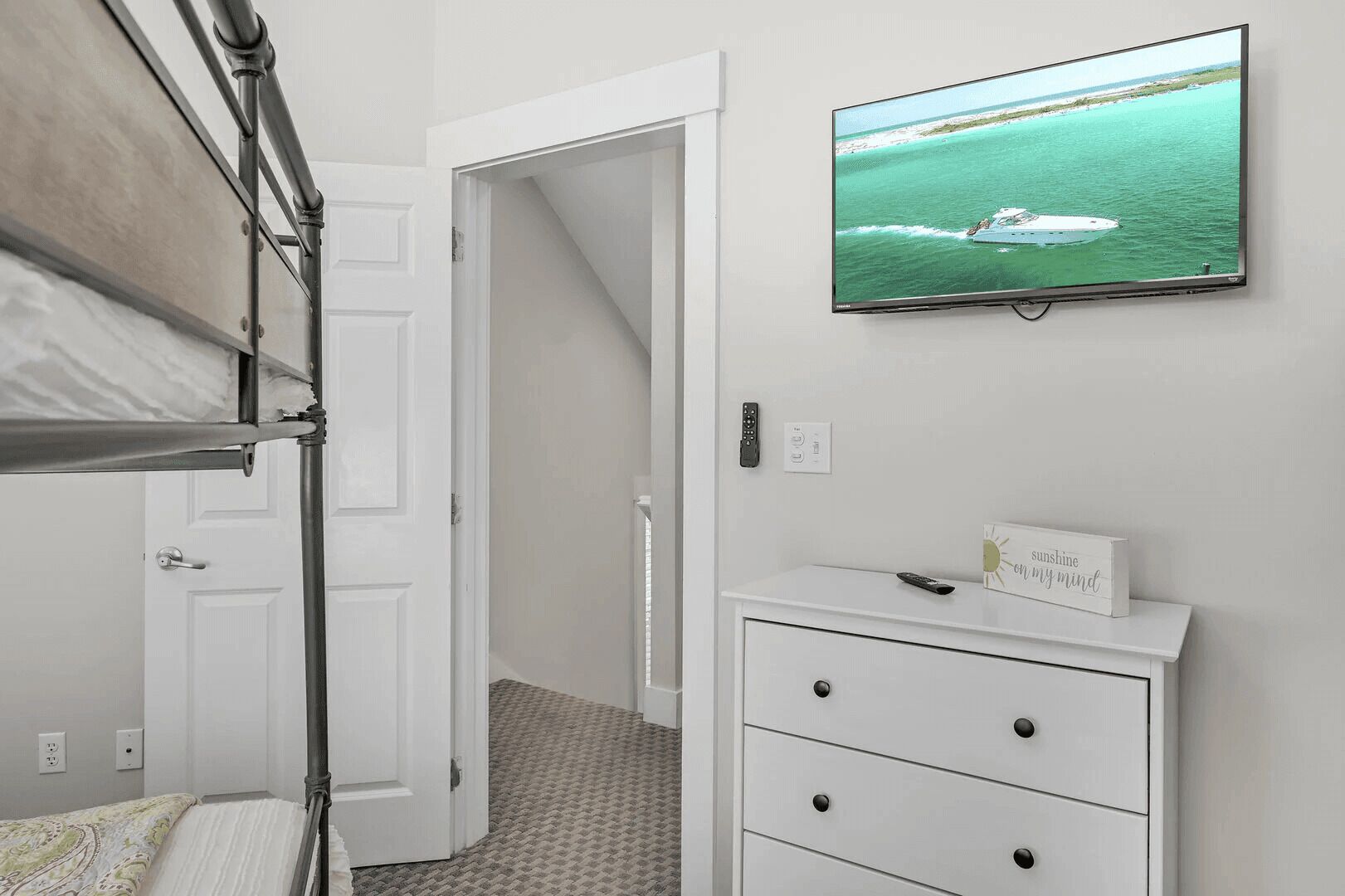 Um quarto com uma cama de solteiro, uma televisão exibindo um barco na água, uma mesa de cabeceira com um controle remoto e um penteado com gavetas.