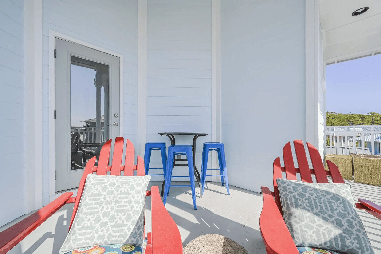 Un espace extérieur abrité avec des chaises Adirondack rouges et des tabourets bleus, une petite table et une porte donnant sur l’intérieur.