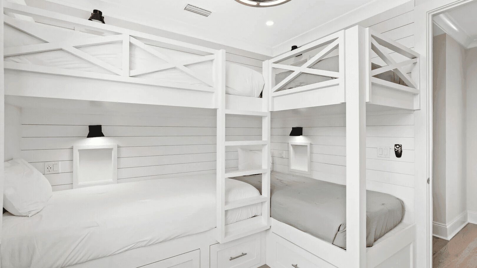 A bunk bed room with white walls, a ceiling fan, and a door leading to another room.