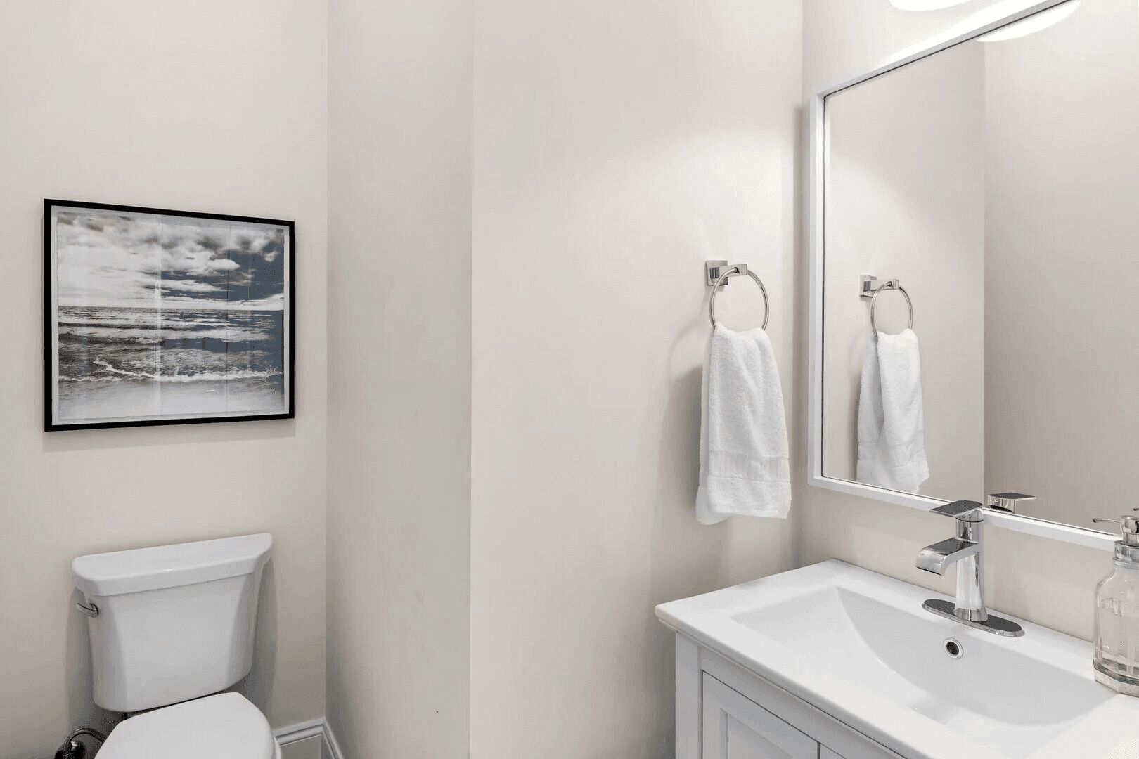 A bathroom with a toilet, a sink, and a mirror. There is a framed picture on the wall.