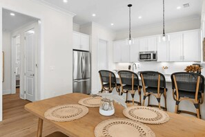 Eine moderne Küche mit weißen Schränken, einem Edelstahlkühlschrank und einer Mikrowelle. Ein Holztisch mit vier Stühlen und zwei gewebten Tischsets steht bereit.