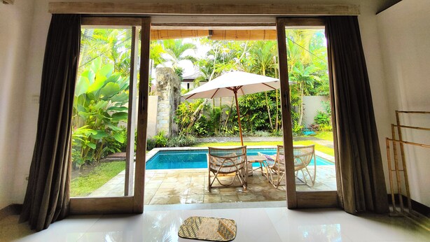 Una habitación con un amplio ventanal corredera que da a un espacio exterior con piscina, mobiliario de patio y exuberante vegetación.