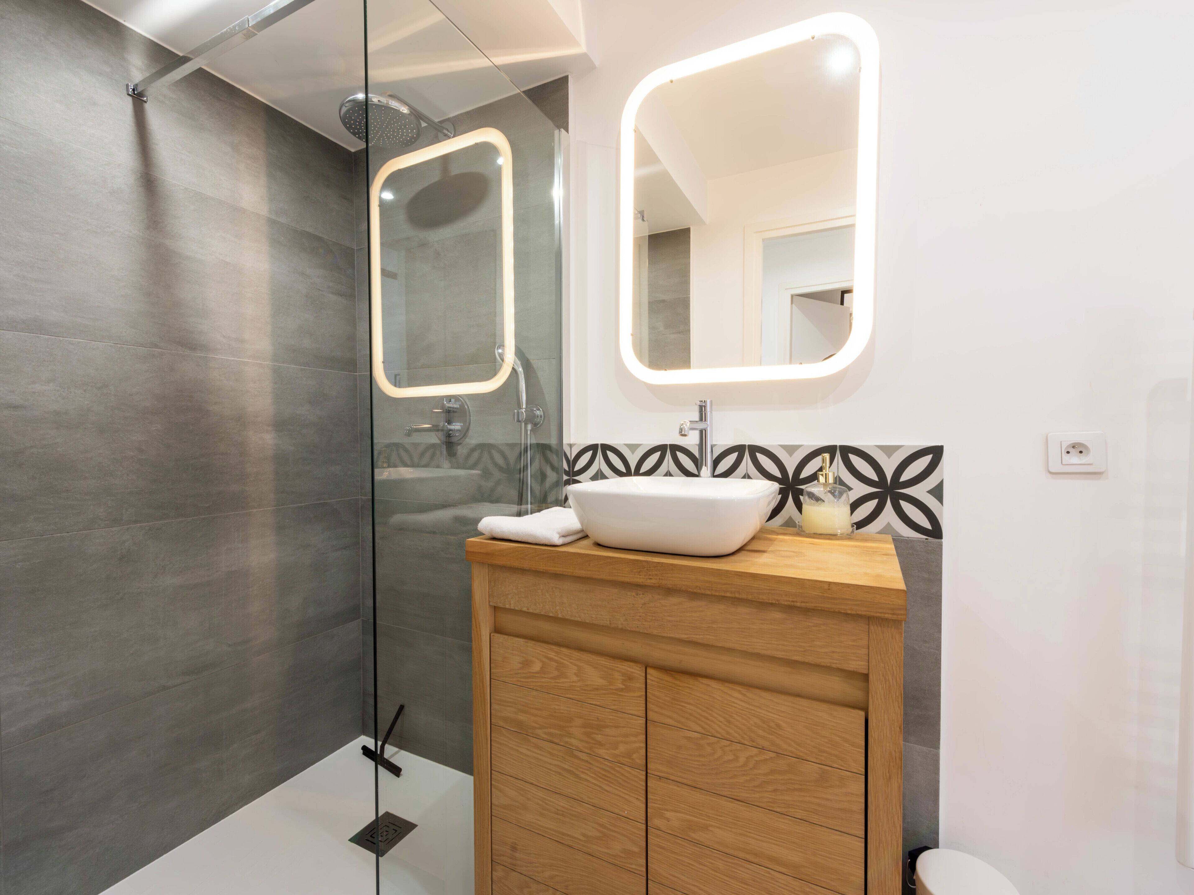 Une salle de bain moderne avec une douche à parois de verre, un meuble-lavabo en bois avec un évier blanc et deux miroirs rétroéclairés.
