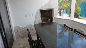 A dining area with a stone wall, a built-in grill, and a view of palm trees. - Czaritza Apart Hotel (Bombinhas)