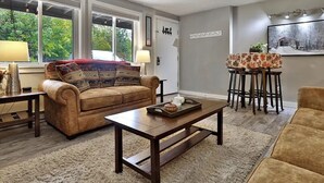 A living room with a sofa, coffee table, and a small table with chairs. There is a window with a view of trees and a door with a light switch. - Killington condo 5 min to killington resort (Killington)