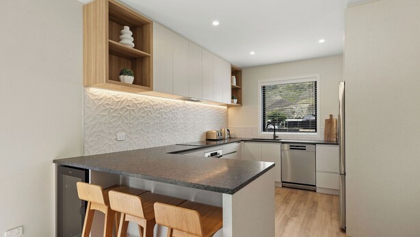 A modern kitchen with a central island, wooden bar stools, built-in shelves, and a view of the outdoors through a window.